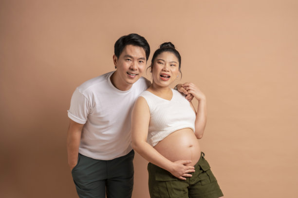 幸福的亚洲情侣家庭照片，孕妇与她的时尚休闲丈夫在米色背景下微笑相拥，洋溢着快乐与幸福的关系。图片下载