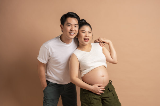 幸福的亚洲情侣家庭照片，孕妇与她的时尚休闲丈夫在米色背景下微笑相拥，洋溢着快乐与幸福的关系。图片下载