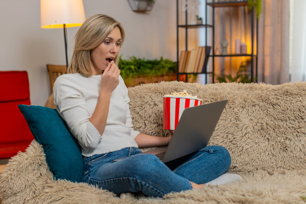 成年女性在家中被震惊的电影时刻惊呆，手里拿着爆米花，盯着笔记本电脑屏幕。图片下载
