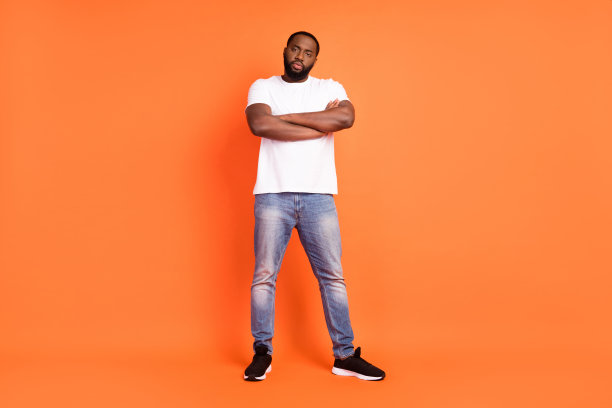 自信严肃的深色皮肤年轻男子双手交叉，站在橙色背景前的全身照片图片下载