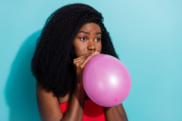 女孩在派对准备中，吸入气球的照片肖像，背景为鲜艳的蓝色。图片下载