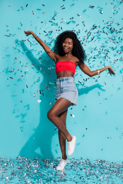 深肤色迷人的年轻女性在蓝色背景下享受好心情，身边飘落着五彩纸屑的全身竖版照片图片下载