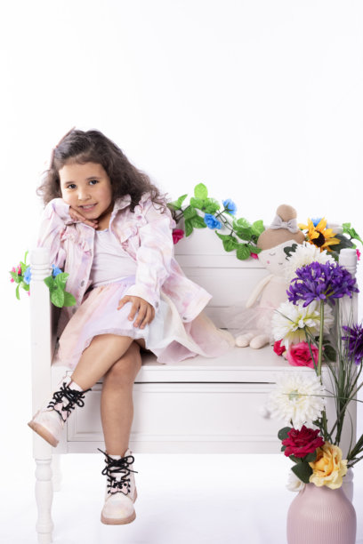 微笑的小女孩坐在装饰精美的白色长椅上，周围有花朵，自信地摆姿势，穿着淡色连衣裙和靴子，展现快乐的童年生活方式照片图片下载