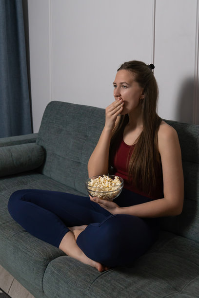 无聊的年轻女性坐在舒适的沙发上，边吃着玻璃碗里的爆米花，边看着一部无趣的电影，感到失望和冷漠。竖版照片图片下载