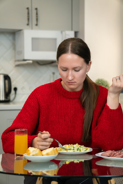 年轻女性穿着红色毛衣，坐在现代厨房的玻璃桌旁，享受着轻食，包括沙拉、香肠、面包和果汁。竖版照片图片下载