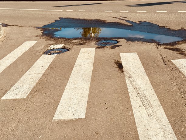 人行横道上有一个大水坑和路面损坏，突显了行人面临的挑战。这张图片展示了人行横道的特写，包含水坑、路面磨损和重要的交通标记。图片下载