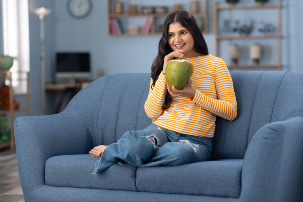 年轻女性穿着现代服装坐在家里的沙发上喝椰子水的照片图片下载