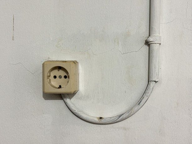简约的镜头展示了一个复古的欧洲电源插座，背景是风化的白色墙壁，旁边是与墙壁颜色相配的连接电缆，暗示着老旧的基础设施和简单的生活方式。图片下载
