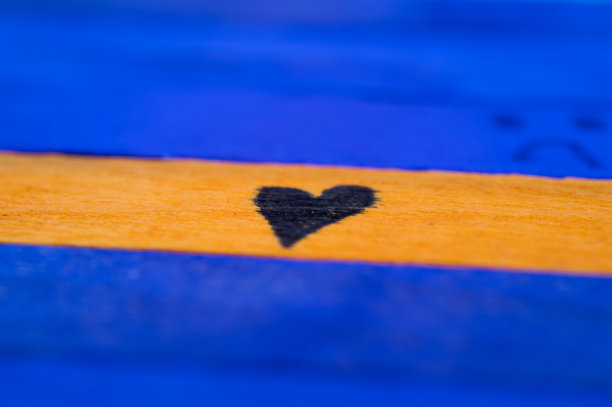 一只手绘的黑色心形图案特写，位于一根黄色木棍上，周围是蓝色木棍，背景模糊的悲伤面孔。图片下载