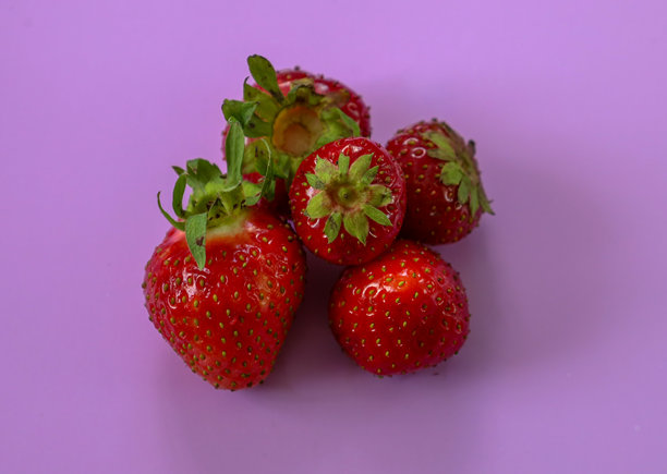 五颗新鲜的红草莓和绿色叶子聚集在统一的紫色背景上。色彩对比使照片生动而吸引人，突显了草莓的自然美。图片下载