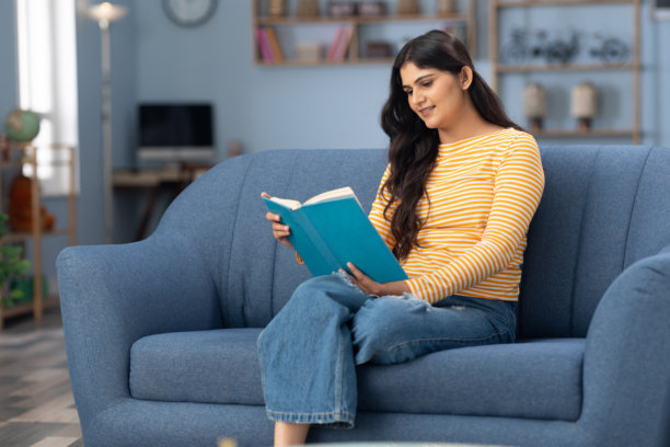 可爱的年轻女孩坐在沙发上阅读书籍，穿着条纹衣服，室内房间的全身照片图片下载