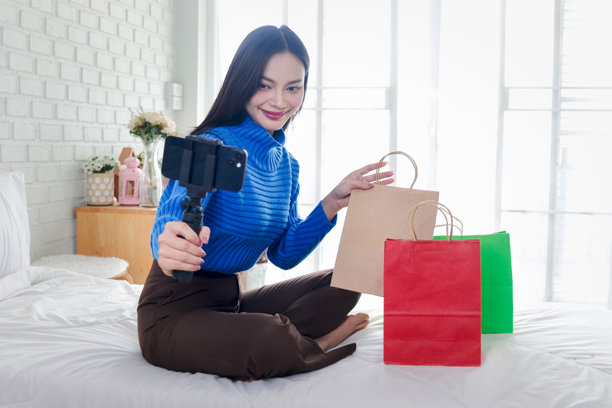 美丽的亚洲时尚影响者女性在卧室里拿着购物袋用智能手机录制社交媒体频道的 vlog。快乐的年轻女性博主正在在线直播播客，拍摄自拍。图片下载