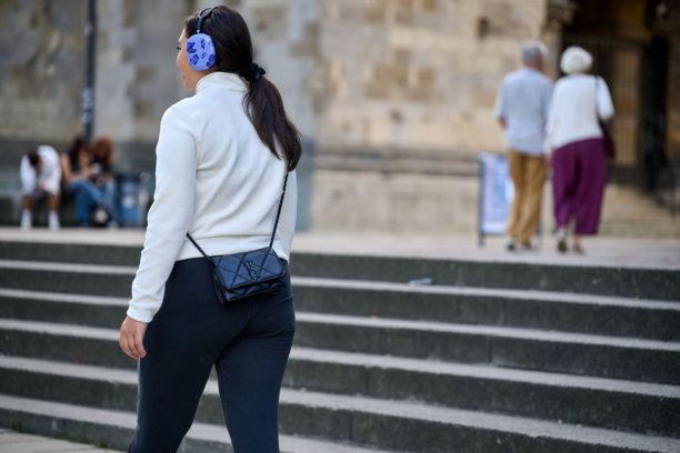 戴着耳机的女性在历史建筑和石阶附近的城市广场上行走，休闲的日间动作场景，适合城市生活和通勤概念的横向街头照片。图片下载