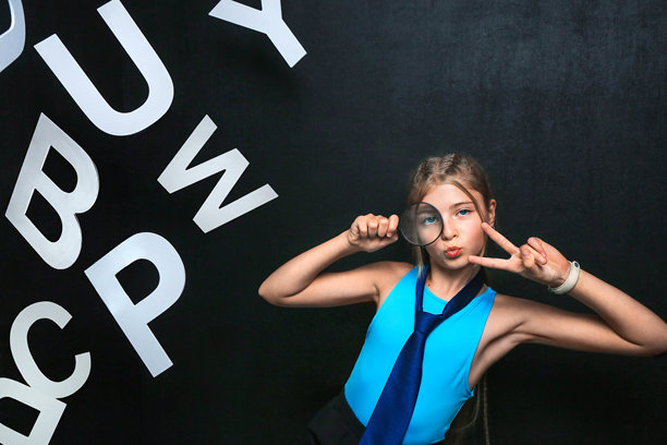 照片中显示一个穿着蓝色上衣和领带的女孩，手持放大镜在脸前，做出和平手势。背景中散落着大白字，营造出一种俏皮和创意的氛围。图片下载