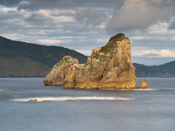 巴斯克海岸宁静的岩石海堆 - 一张长时间曝光的照片，展示了海中央的大型岩石形成，水面平滑，背景是起伏的山脉。图片下载