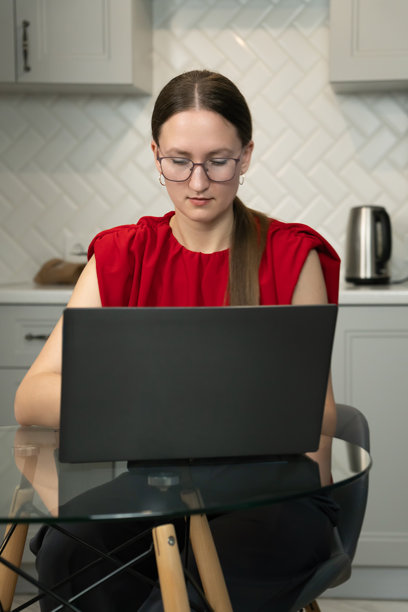 专注的女商人戴着眼镜，穿着红色上衣，专心致志地在厨房桌子上使用笔记本电脑远程工作，体现了现代自由职业的生活。竖版照片图片下载