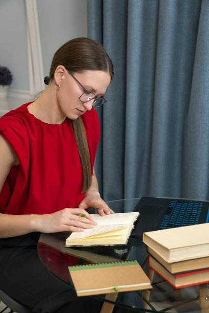 穿着红色上衣的女性专注于笔记本电脑，分析数据。她坐在玻璃桌旁，周围是书籍，身处明亮现代的房间，面带沉思的表情。竖版照片图片下载