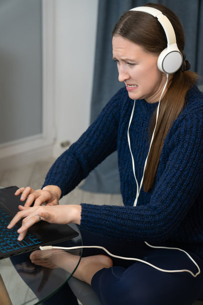 年轻女性戴着耳机在家工作，坐在椅子上，双腿交叉，正在键盘上打字并看着屏幕。竖版照片图片下载