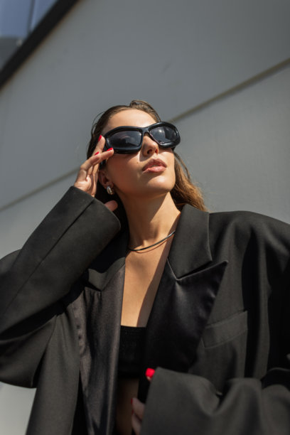 年轻女性模特穿着时尚的宽大外套和配饰，在阳光下的户外摆姿势。酷炫的美丽女孩在城市中戴上黑色衣服和太阳镜。图片下载