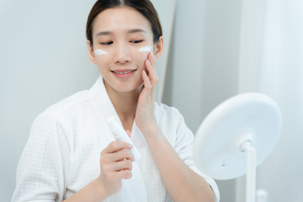 美丽的亚洲女性坐在镜子前，健康的面容正在涂抹面霜和化妆品。皮肤霜、抗皱、婴儿般的面孔、美白、保湿、收缩毛孔精华的广告。图片下载