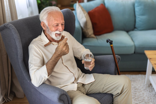 成熟男性的晨间例行 - 手持药物和水。照片中是一位老年男性在家中拿着药丸和水杯。老人坐在扶手椅上，正在经历抑郁。男子服用含有Omega-3的药丸，手中握着一杯新鲜水。图片下载