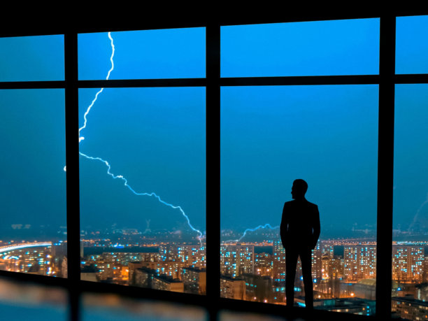 身穿西装的商务人士剪影站在窗边，窗外是城市的暴风雨氛围，夜晚高楼大厦的办公室内闪烁着灯光。图片下载