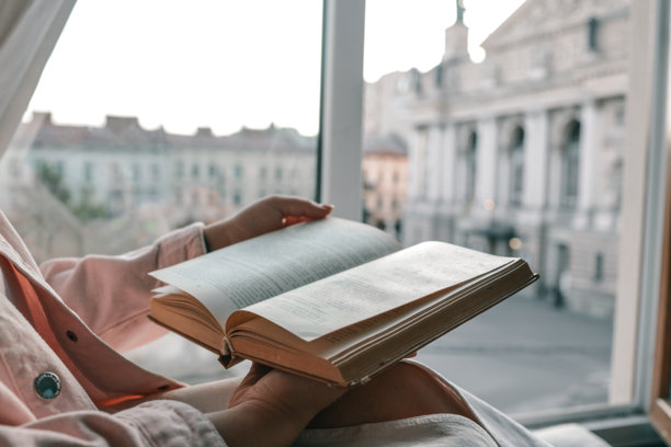一个人在窗边拿着一本打开的复古书籍，窗外可以看到历史悠久的欧洲广场和歌剧院，营造出一个在日落时分安静阅读和反思的美好时刻。图片下载