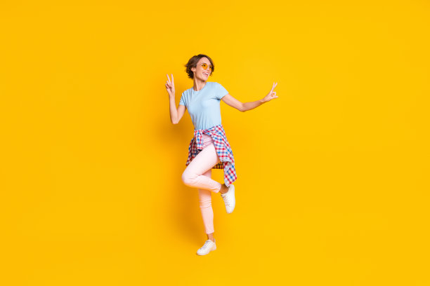 全身肖像照片，女性一条腿站立，跳舞时做出两个V字手势，穿着紧身腰部衬衫，背景为鲜艳的黄色。图片下载