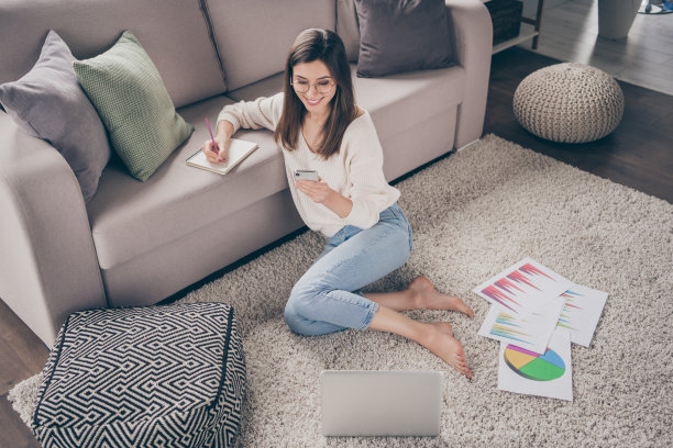 高角度全景照片，快乐迷人的女孩坐在地板上，穿着牛仔裤，使用手机写笔记，进行远程学习，室内环境。图片下载