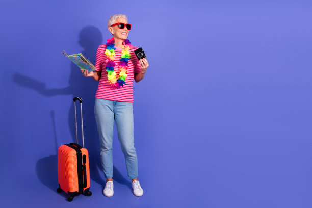 一位老年女性手持地图和相机，旁边放着行李箱，背景为紫色，适合旅行和生活方式的库存图片。图片下载