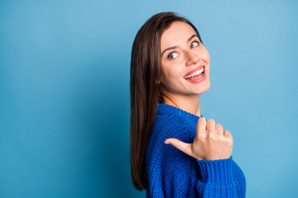 年轻快乐兴奋的可爱女孩侧面照片，微笑着用拇指指向空白处，背景为蓝色。图片下载
