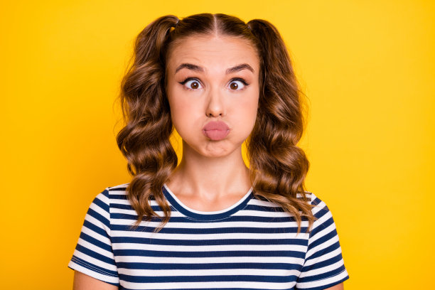 穿着条纹服装、撅嘴、脸颊鼓鼓的可爱搞笑年轻女士的照片，背景为黄色。图片下载