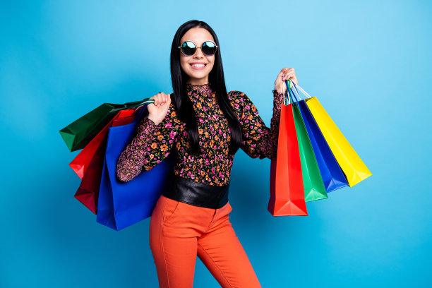 酷女孩手持多只购物袋，佩戴复古眼镜，穿着花卉印花衬衫，背景为蓝色。图片下载