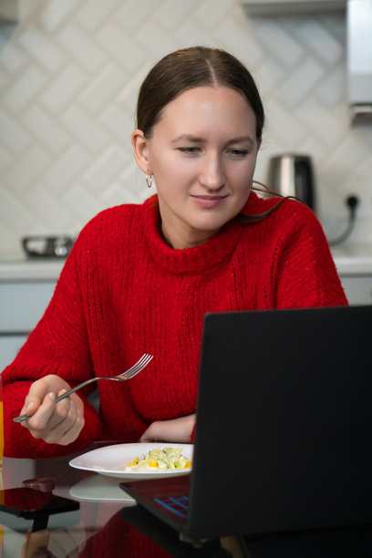 年轻女性穿着鲜艳的红色毛衣，在时尚现代的厨房里专注于笔记本电脑，享用健康沙拉，融合了科技与健康的用餐体验。竖版照片图片下载