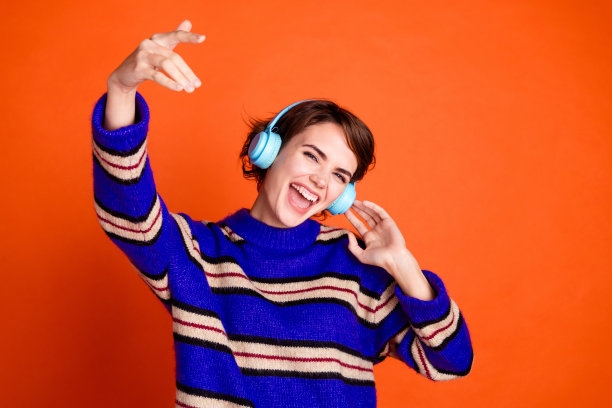 一位棕发的千禧女性模特穿着条纹毛衣，享受着耳机，独自在橙色背景上跳舞的照片图片下载