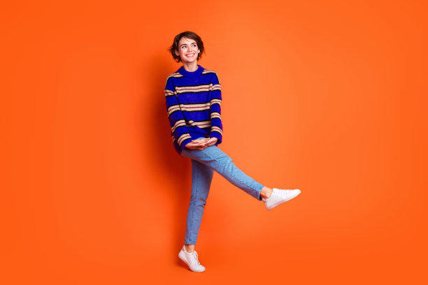 年轻女孩穿着蓝色条纹毛衣和牛仔裤，开心地在橙色背景上跳舞的全身照片。图片下载