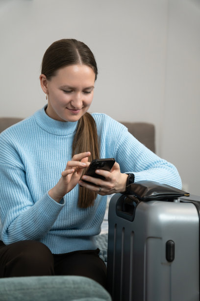 年轻女性坐在床上，旁边放着打包好的行李箱，使用智能手机规划旅行并进行在线预订或查看旅行信息。竖版照片图片下载