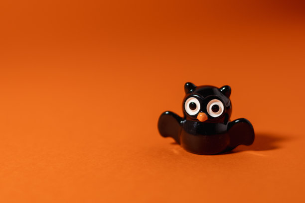 小猫头鹰雕像特写，拥有大眼睛，在橙色背景上投射出戏剧性的蝙蝠形状阴影。这个充满趣味的万圣节灵感布置营造出一种既神秘又可爱的氛围，非常适合季节性广告和儿童派对邀请函。图片下载