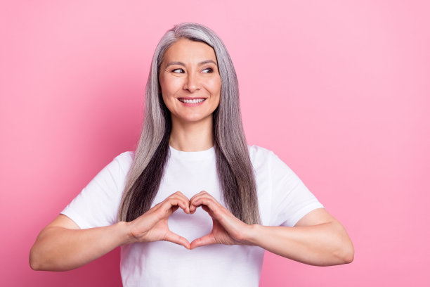 一位银发老妇人在粉色背景前用手指做出心形手势的肖像照片图片下载