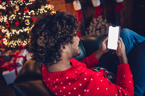 侧面照片，放松的男士躺在沙发上，手持智能手机，利用空白空间预订圣诞假期的度假酒店。图片下载