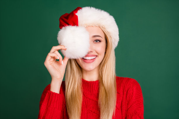 戴着圣诞帽的金发女性在绿色背景下用毛皮遮住一只眼睛的肖像照片图片下载