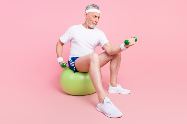全身照片：一名男子坐在健身球上，手持哑铃进行锻炼，背景为柔和的粉色。图片下载