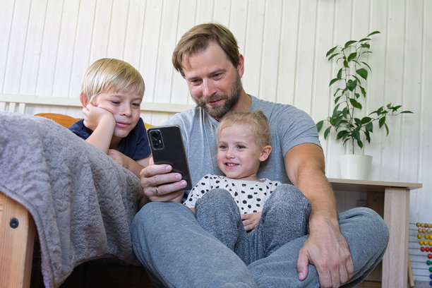 年轻爸爸和孩子们一起使用智能手机。男人在手机上观看内容。快乐的父亲、女儿和儿子在看搞笑视频时开心地微笑，假期的照片，玩手机游戏。图片下载