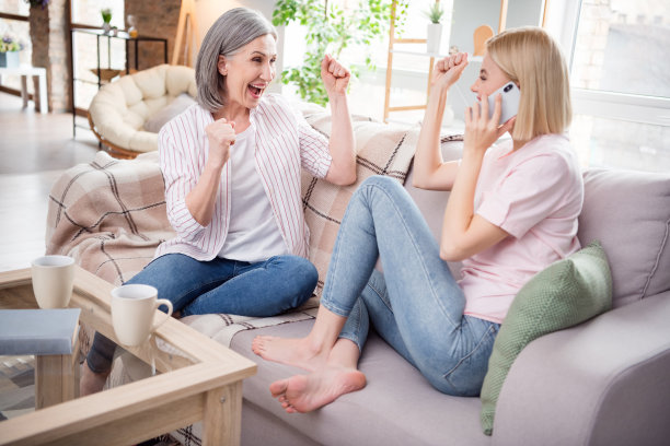全身照片，灰金色头发的女性家庭成员庆祝，坐在沙发上，手持手机，室内家中。图片下载
