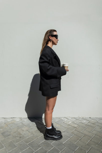时尚的年轻女性模特穿着潮流黑色服装，戴着酷炫的眼镜，脚踩靴子，手里拿着外带咖啡杯，站在城市的灰色墙壁旁。漂亮的魅力商务女孩。图片下载