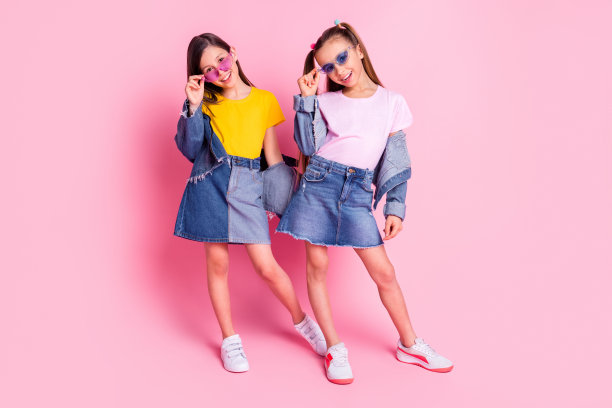 乐观的棕发和金发女孩穿着黄色和粉色的T恤裙，背景为粉色，照片为全身照。图片下载