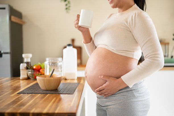 孕妇在厨房里站着，手里拿着杯子喝水，轻抚着肚子。图片下载