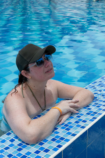 一位戴着黑色棒球帽和太阳镜的宁静女性在碧绿色的泳池中放松。这张图片捕捉了独自旅行中的宁静时刻，反映出一种平和而宁静的度假生活方式。图片下载