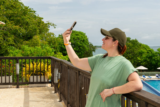 一位穿着绿色连衣裙和棒球帽的快乐女性在阳台上用智能手机自拍。这张图片反映了数字游牧生活方式，非常适合旅游和科技概念。图片下载