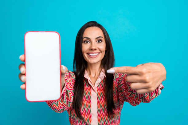 漂亮年轻女孩的照片肖像，惊讶地拿着显示白色屏幕的手机，穿着时尚印花服装，背景为青色。图片下载
